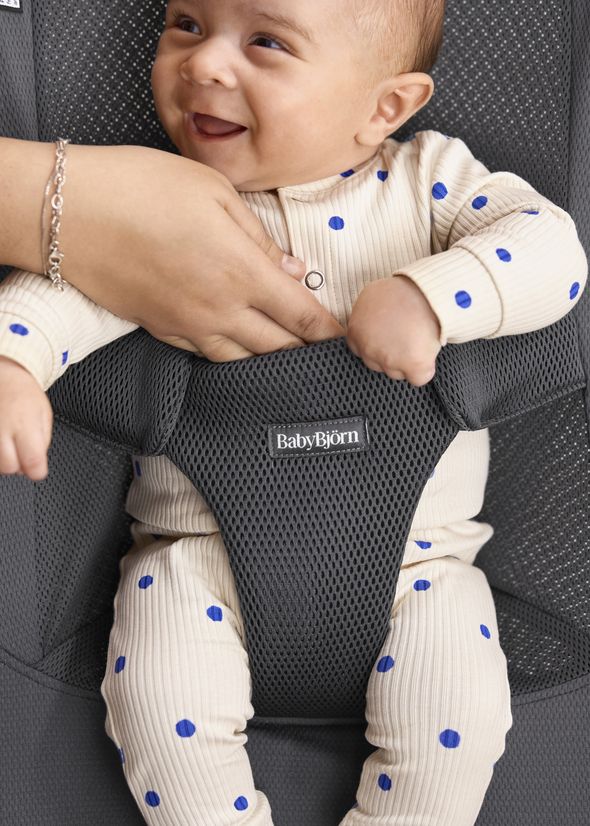 Smiling baby in a beige onesie with blue dots, secured in a black BabyBjörn bouncer.