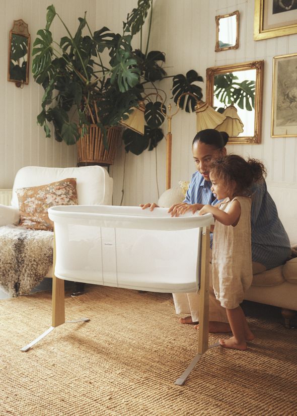 A woman and child stand beside a modern white bassinet in a cozy room with large green plants and ornate wall mirrors.
