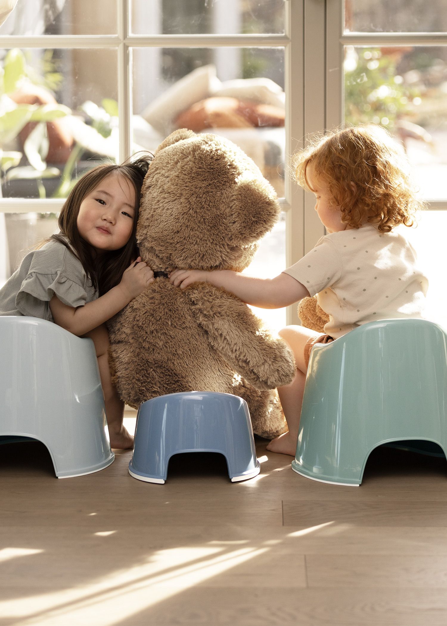 Two children sit on stools, hugging a large teddy bear in a sunlit room with a glass door.