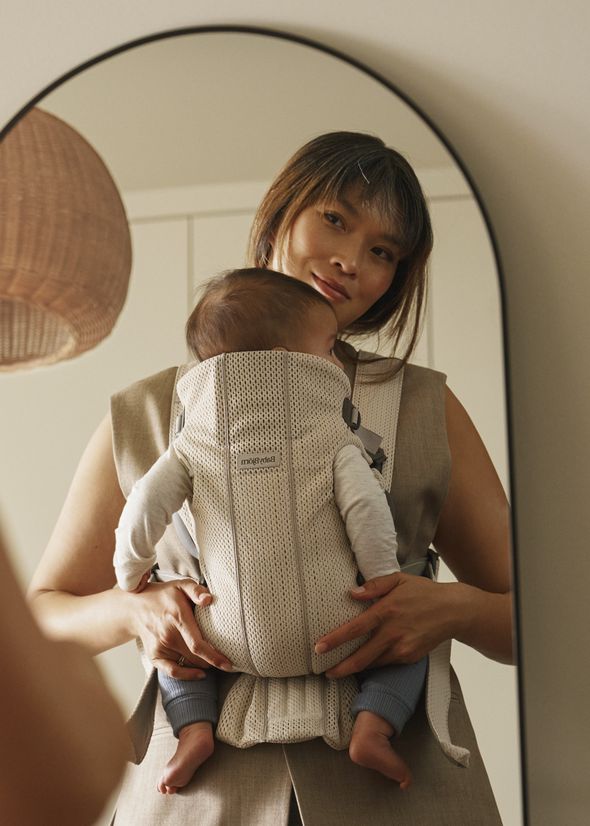 Woman holds baby in a beige carrier, reflecting in a mirror. She wears a tan sleeveless outfit, with a wicker lampshade in the background.