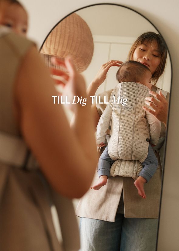 Mother holding baby in a front carrier, standing in front of a mirror. Text on the mirror reads "TILL Dig TILL Mig."