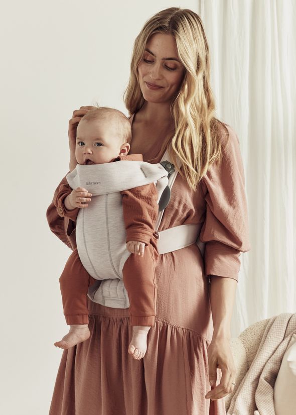 A woman with long hair wearing a pink dress smiles while holding a baby in a gray BabyBjörn carrier.