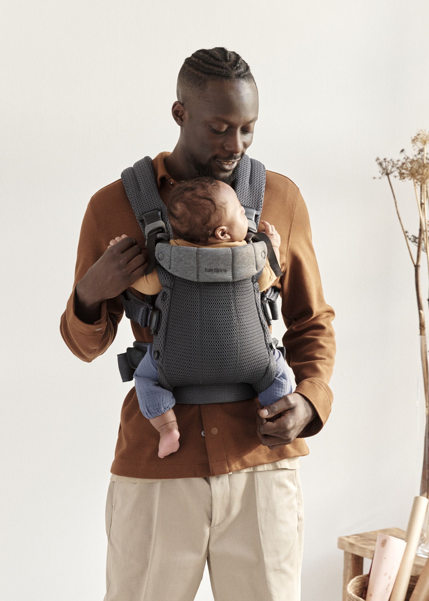 Un homme souriant porte un bébé endormi dans un porte-bébé gris, se tenant debout dans une pièce lumineuse avec des plantes en arrière-plan.