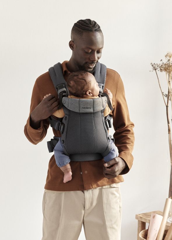A man with short hair and a beard wears an orange shirt and holds a baby in a gray baby carrier. The baby is sleeping against his chest.