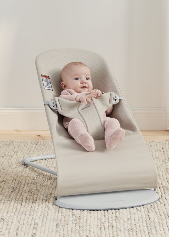 A baby sits comfortably in a beige baby bouncer on a carpeted floor, wearing a pink outfit and looking content.