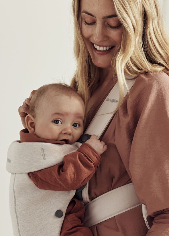 A smiling woman holds a baby in a beige carrier. The baby looks curiously at the camera, wearing a rust-colored outfit.