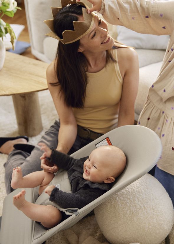 A smiling woman in a crown looks at a laughing baby in a bouncer, while another person adjusts the crown in a cozy room.