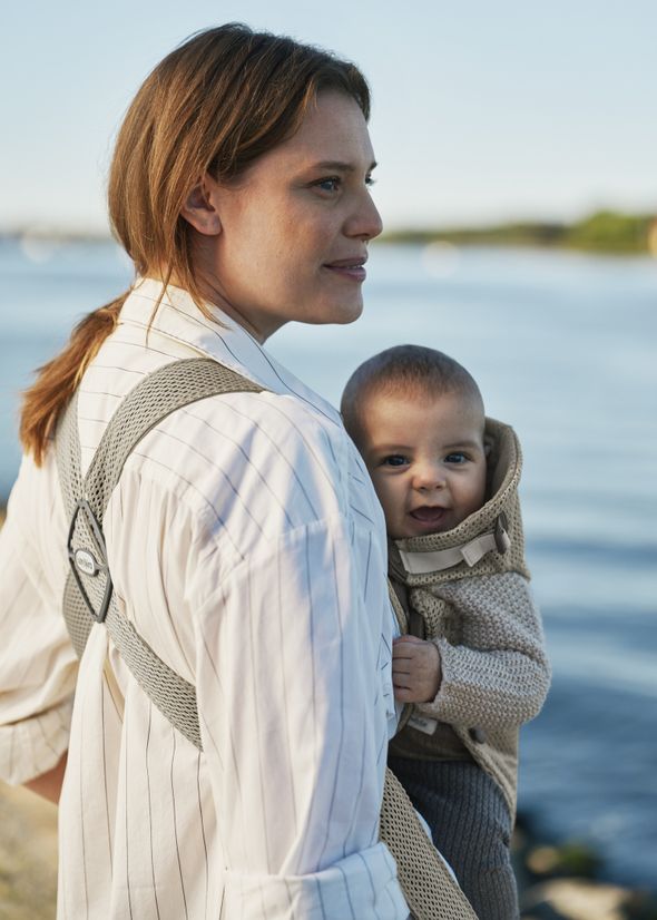 Woman with a baby in a carrier standing by a calm lake, both looking content under a clear blue sky.