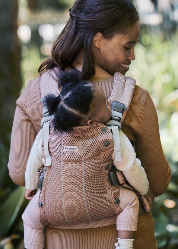 Woman wearing a tan sweater carries a sleeping baby on her back in a pink mesh baby carrier outdoors.