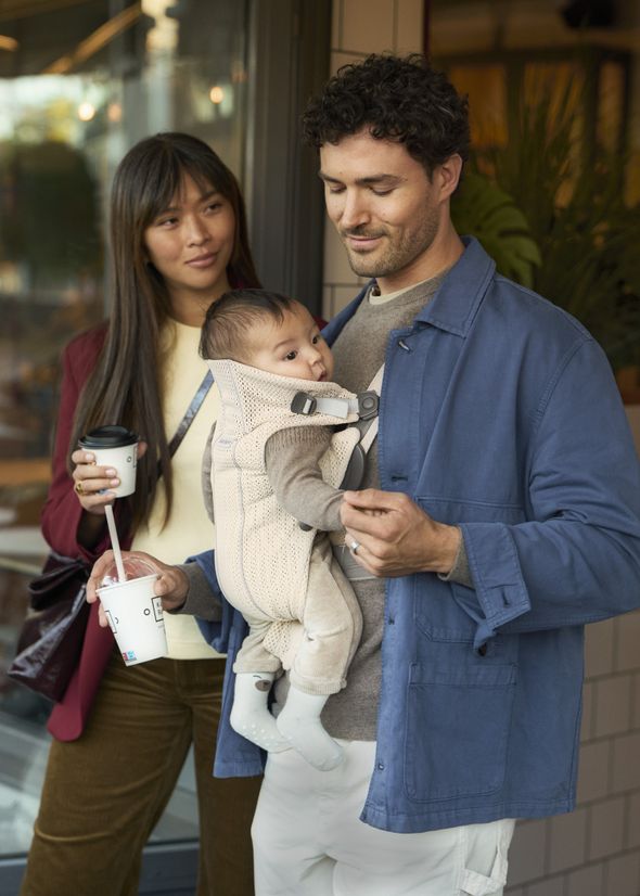 A man carries a baby in a cream coloured carrier while a woman holds coffee. They are outside a building, smiling and casually dressed.