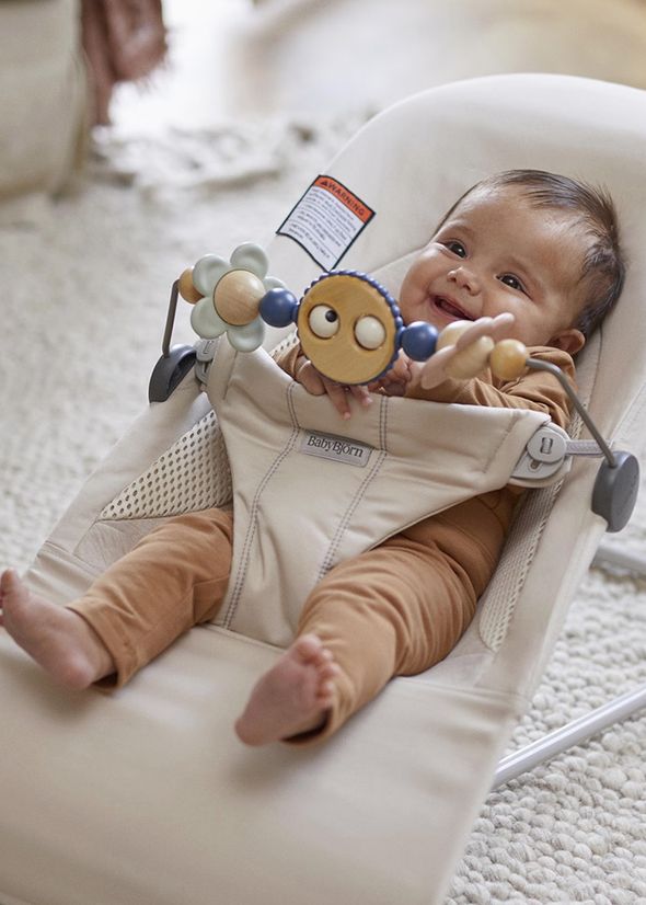 A smiling baby in a beige bouncer with a toy bar, wearing a brown outfit, lies on a patterned carpet.