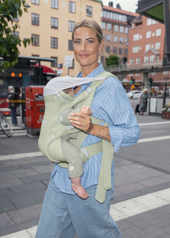 A person with blond hair, wearing a blue striped shirt and jeans, carries a baby in a green baby carrier on a city street.