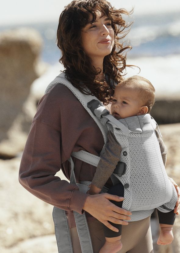 Woman with long hair carrying a baby in a gray carrier, standing on a beach with rocks and ocean in the background.