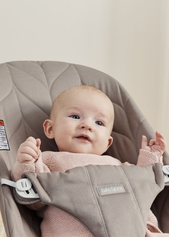 Baby in a beige bouncer wearing a pink sweater, looking up with a curious expression.