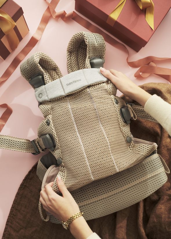 Hands adjusting a beige baby carrier with mesh fabric on a pink surface, surrounded by gift boxes and ribbons.