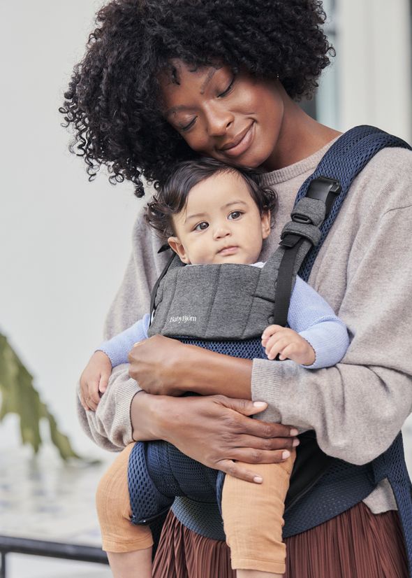 A smiling woman holds a baby in a gray and blue carrier, gently leaning her head on the baby's head. The baby looks curiously at the camera.