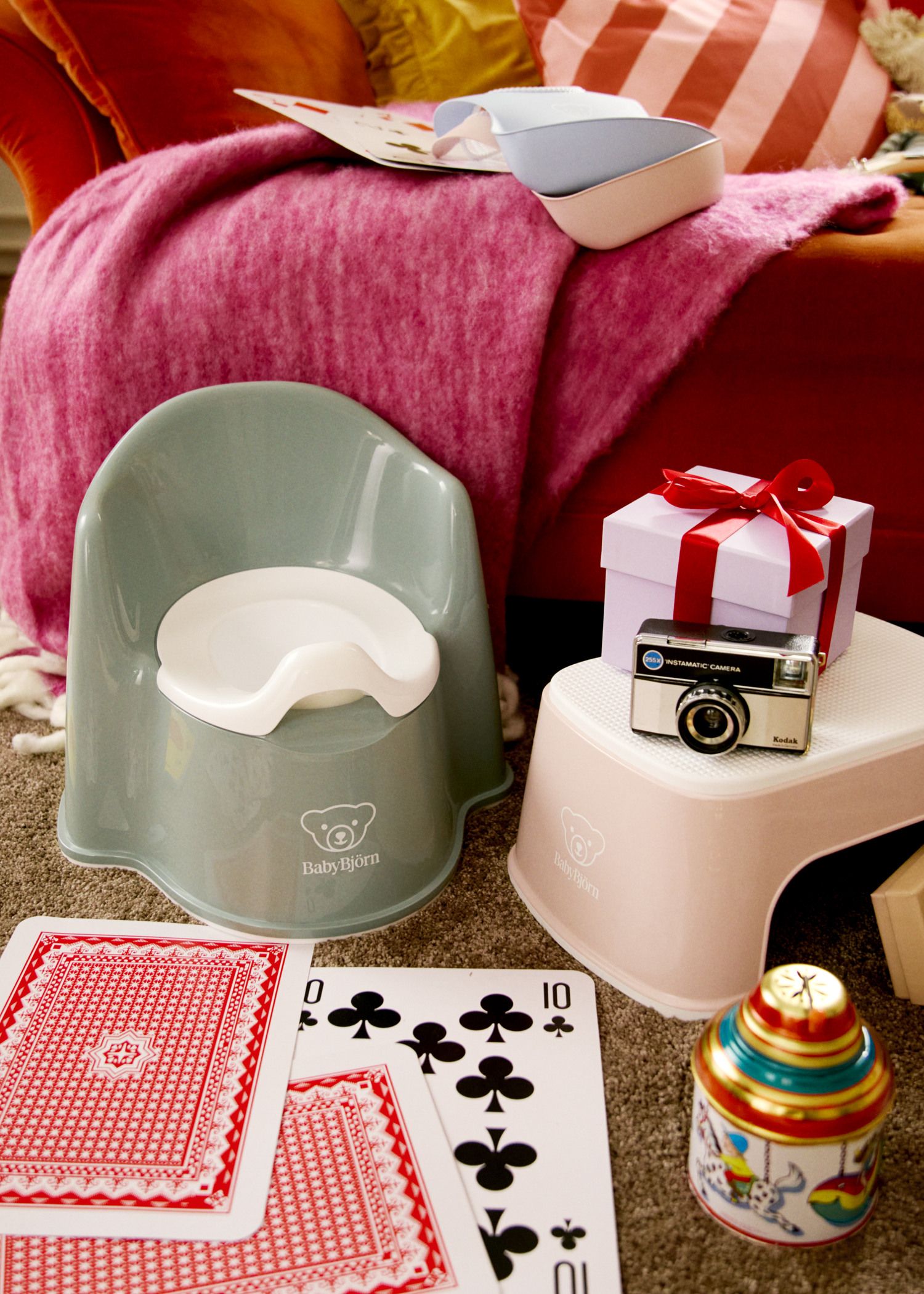 Children's potty and step stool on a carpet with playing cards, a toy camera, and a gift box. A pink blanket is draped over furniture.