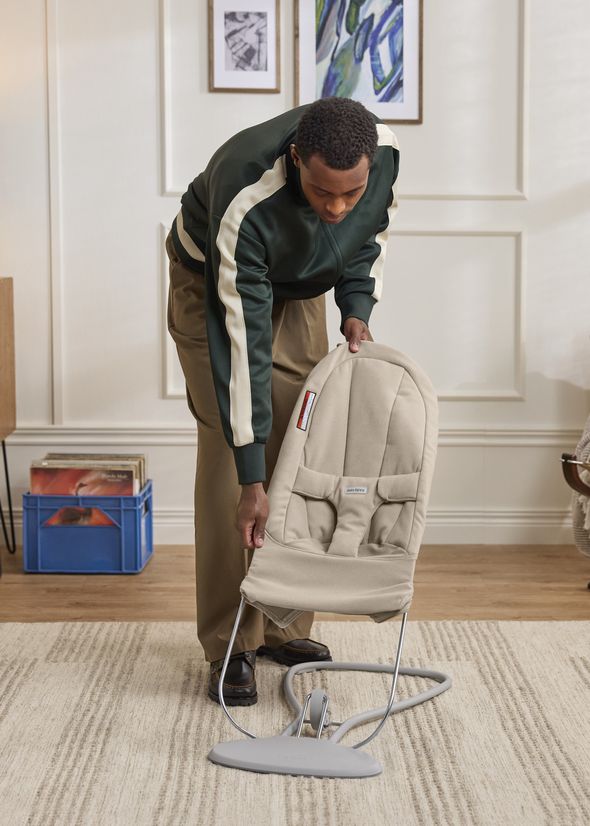 A person in a green jacket sets up a beige baby bouncer in a cozy, decorated room with art on the walls.