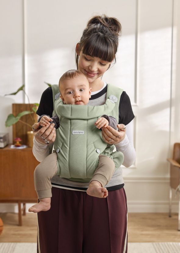 A woman smiles while carrying a baby in a green baby carrier indoors, with a plant and wooden furniture in the background.