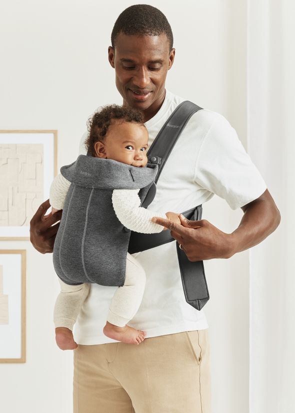 A man smiling at a baby in a gray carrier, both in a bright room with white walls and beige decor.