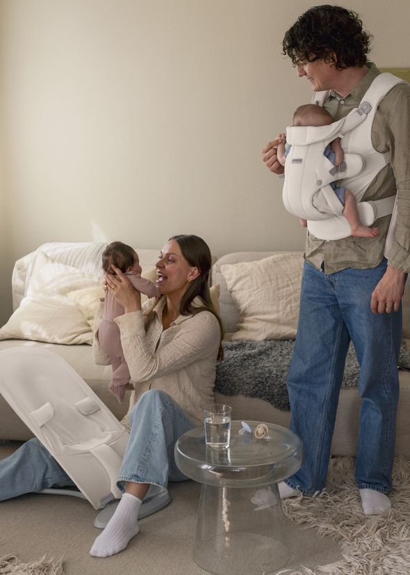 A man holds a baby in a carrier, while a woman plays with another baby on a couch. A glass table with a pacifier is in the foreground.