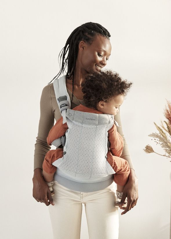 A woman smiling, holding a child in a light gray baby carrier. The child is wearing an orange outfit and looking down. Neutral background.