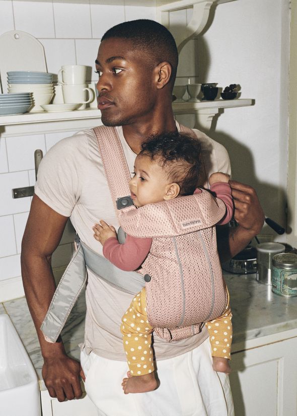 Man in a kitchen wearing a light shirt, holding a baby in a pink carrier. Shelves with dishes are visible in the background.