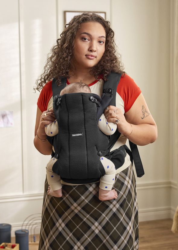 A woman stands indoors, holding a baby in a black carrier. The baby wears a dotted onesie, and the room has neutral decor.