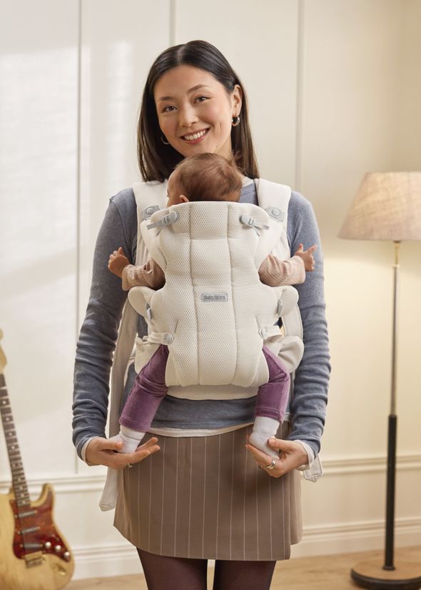 A woman smiling while carrying a baby in a white front carrier, standing in a cozy room with a floor lamp and guitar in the background.