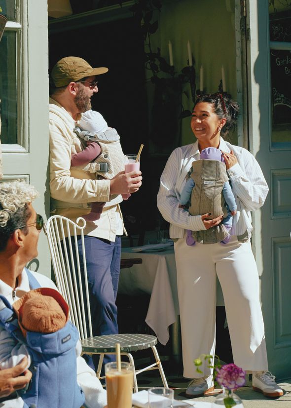A man and woman, each holding a baby in a carrier, smile and chat outside a café. Another person sits nearby, also with a baby.