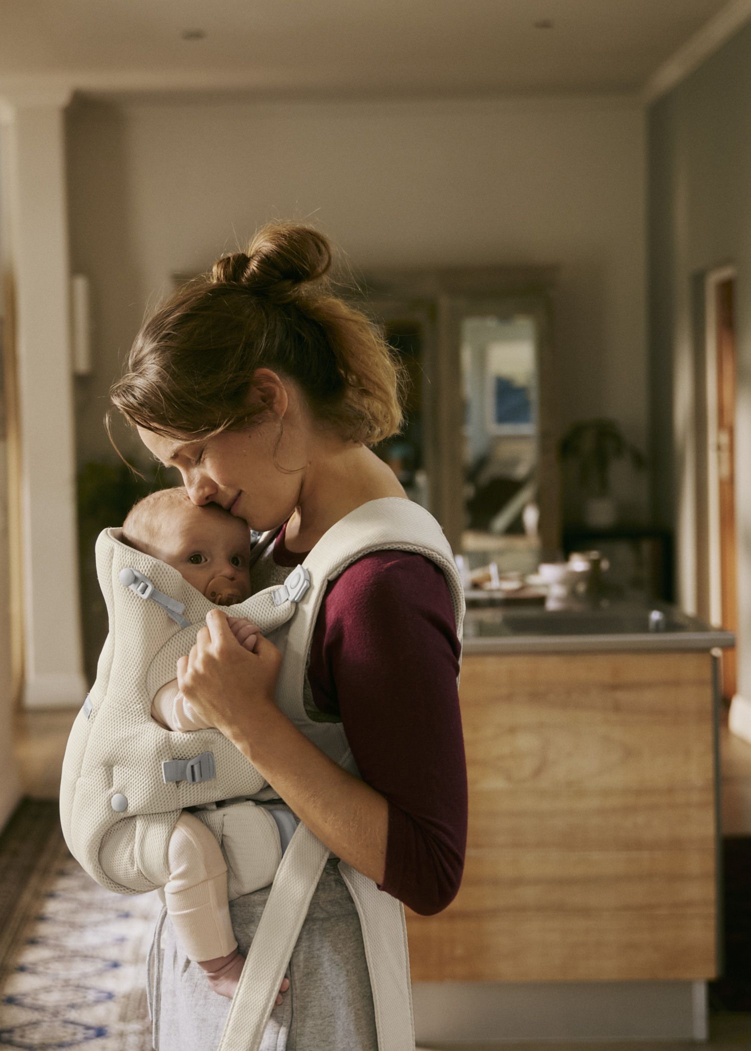 Mother holding a baby in a gray carrier, softly kissing the child's head, in a warmly lit room.