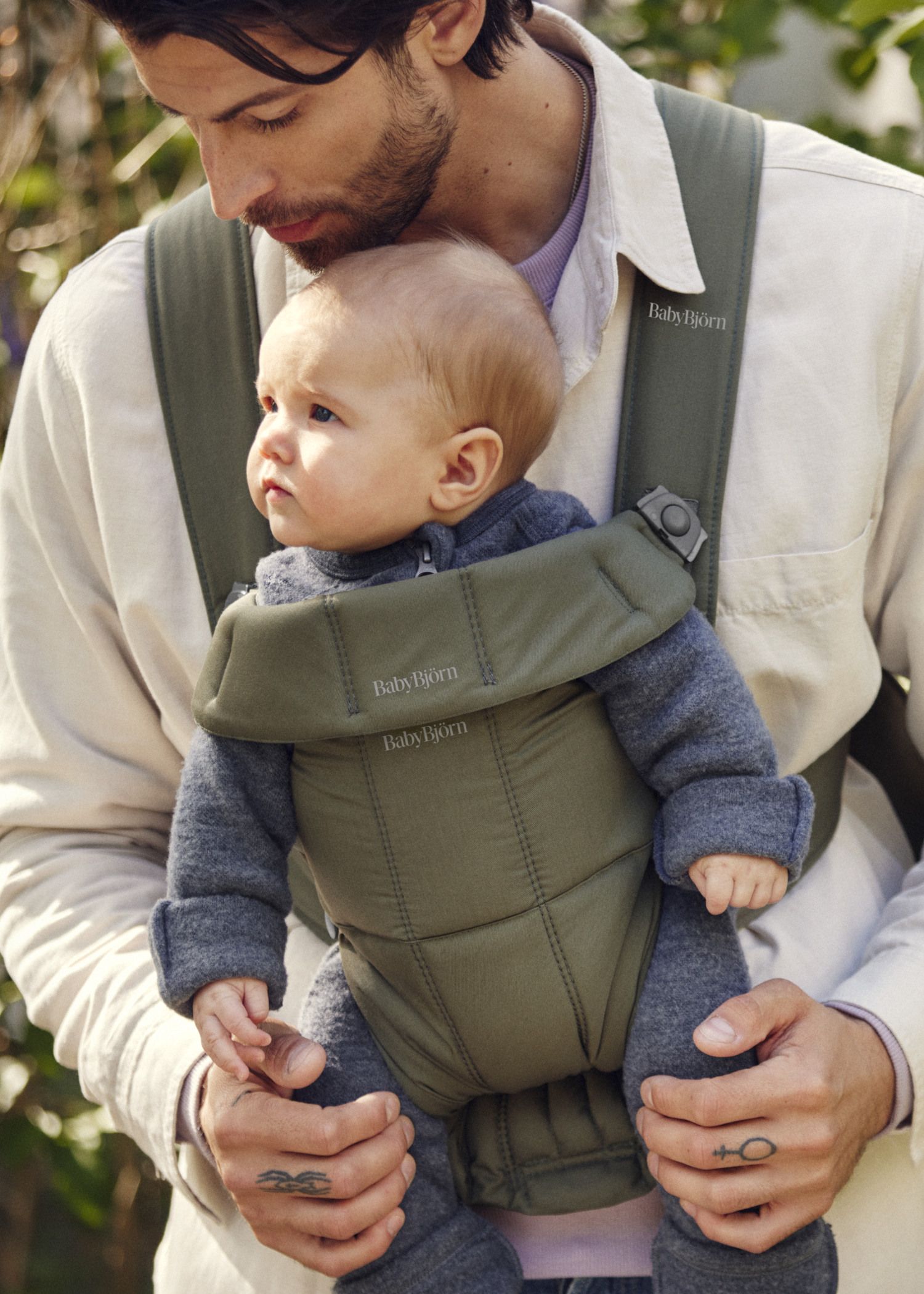 A man holds a baby in an olive green carrier. The baby, wearing a gray outfit, looks intently to the side. The man has a gentle expression.