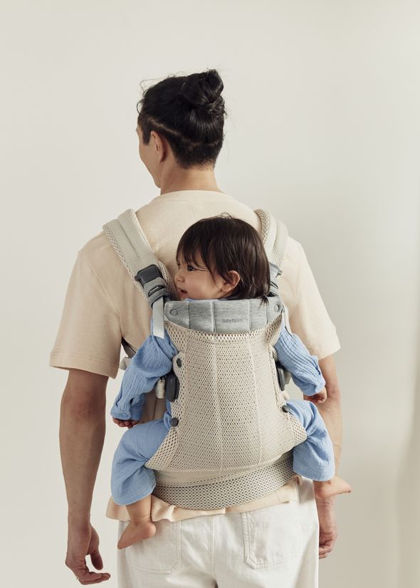 An adult wearing a beige baby carrier holds a child dressed in blue, secured to their back. The adult is facing away, standing against a light background.