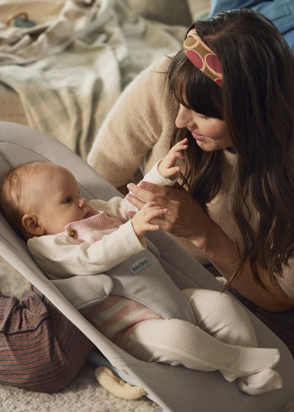 A woman smiles at a baby lying in a gray bouncer. The baby reaches out, wearing a white and pink outfit. Cozy atmosphere.