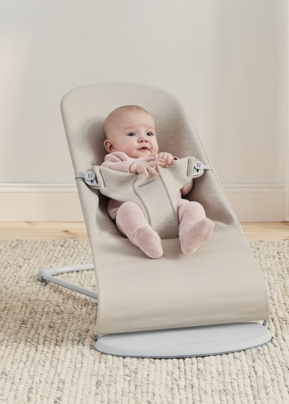 A baby in a pink outfit sits in a beige baby bouncer on a light-colored carpet.