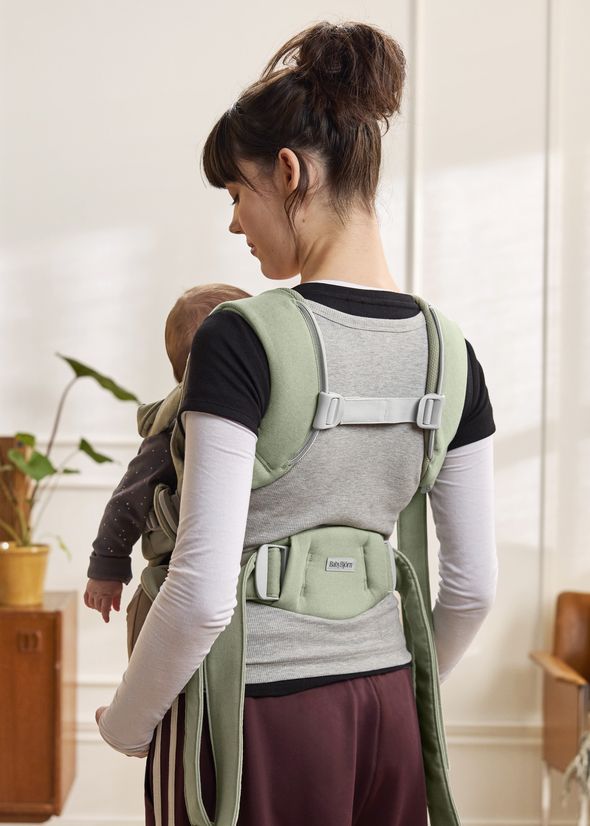 Woman with hair in a bun carries a baby in a green baby carrier, standing in a room with plants and light-colored walls.