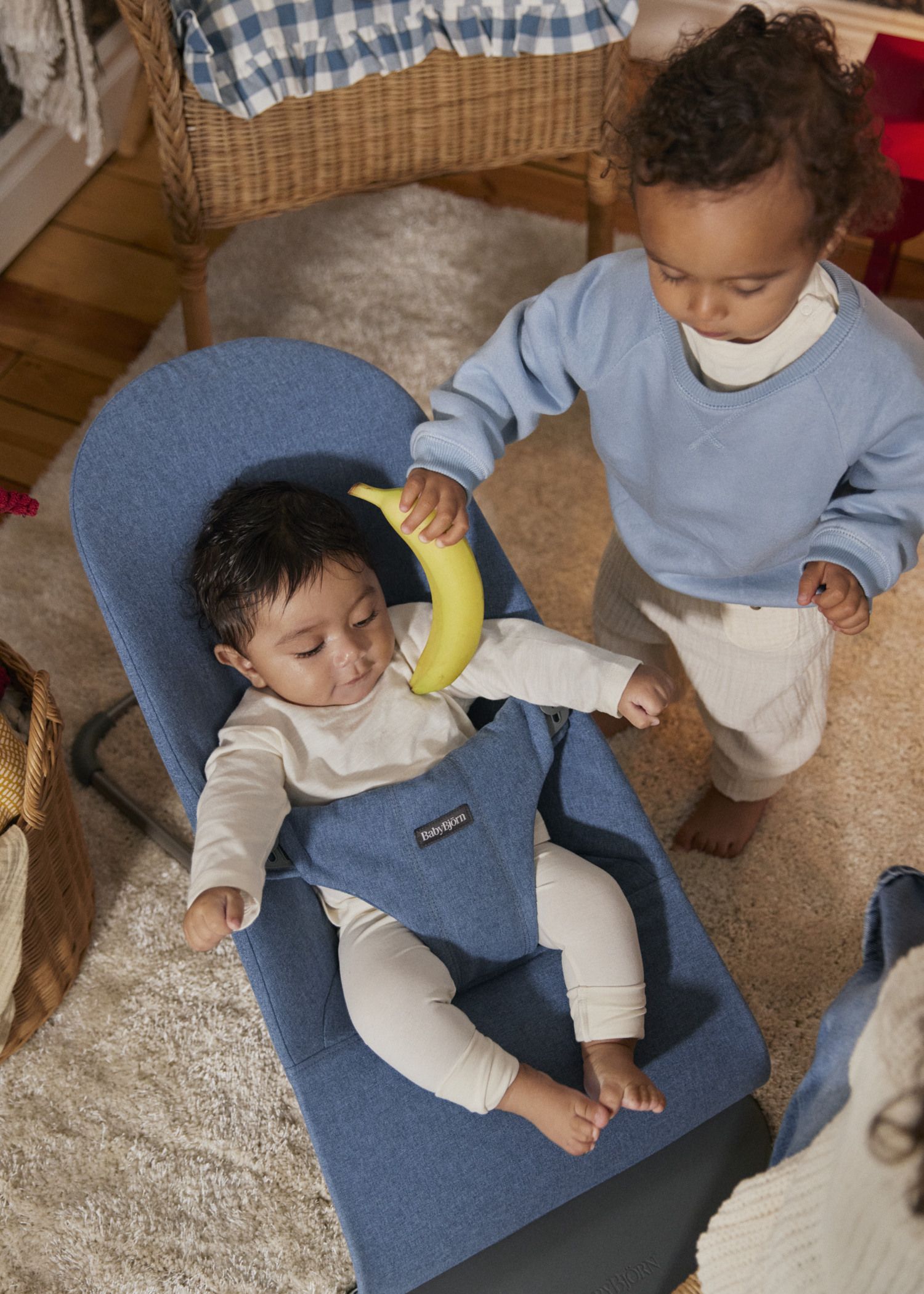 A toddler in a blue sweater playfully holds a banana over a baby in a blue bouncer seat. The room has a cozy, carpeted floor and wicker baskets.