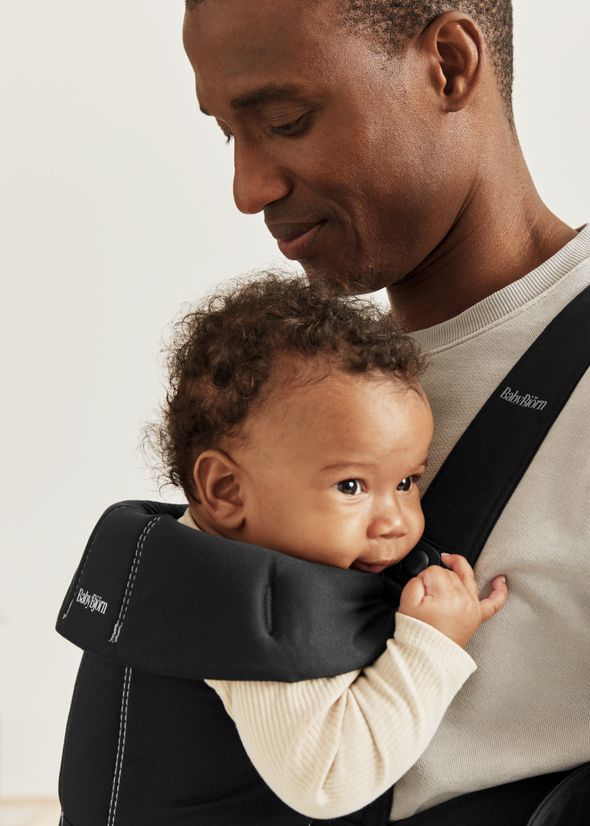 A man gently holds a baby in a front carrier, looking down at the child with a warm expression. The baby is comfortably nestled and sucking their thumb.