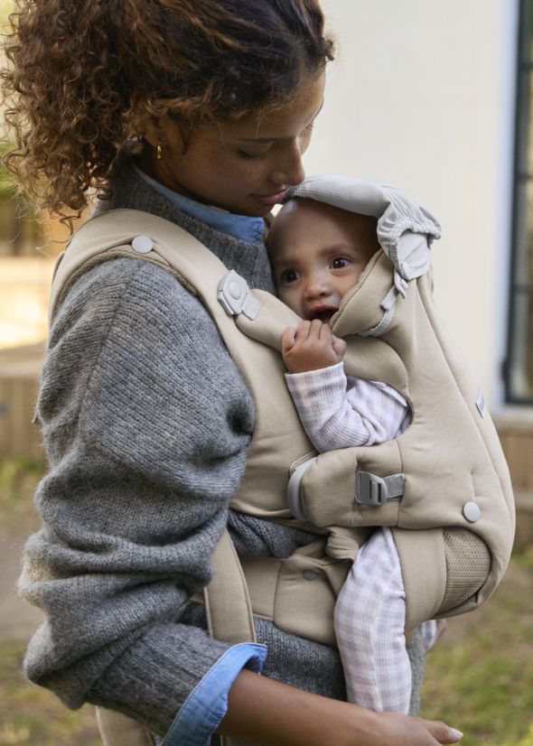 Woman holding a baby in a beige carrier, wearing a gray sweater. The baby looks cozy and rested, outdoors with soft natural light.