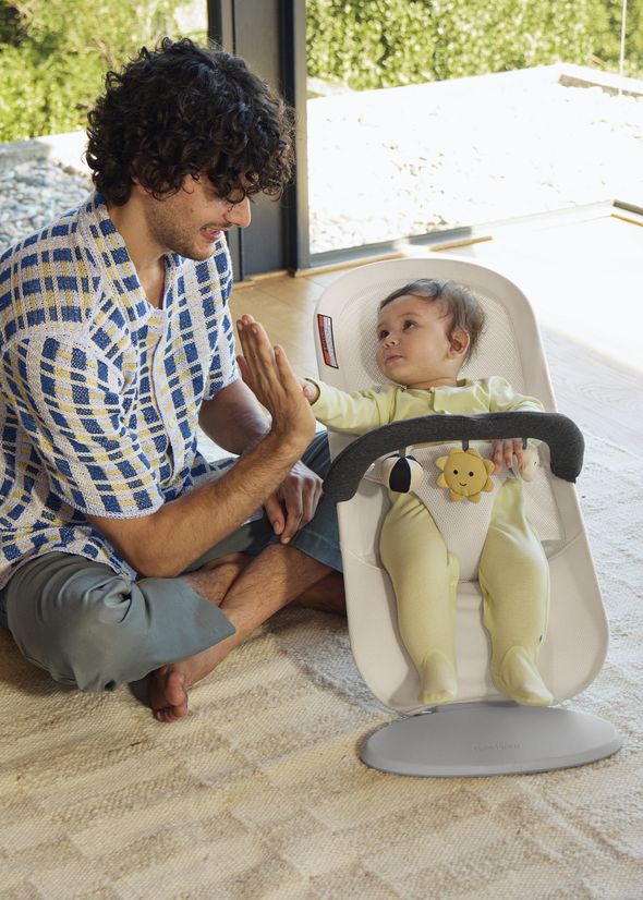 A man playfully gives a high-five to a baby sitting in a bouncer, surrounded by natural light from a large window.
