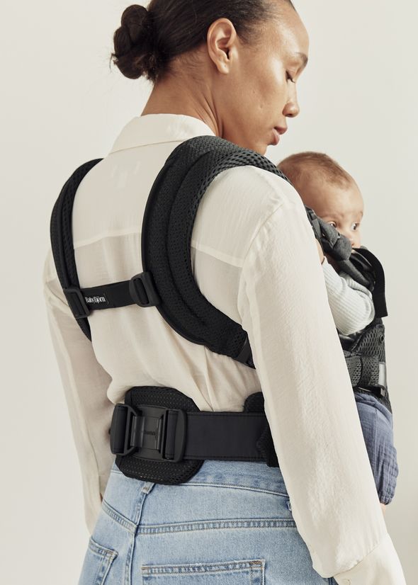 A woman wearing a white shirt and denim jeans carries a baby in a black front-facing baby carrier, looking calmly to one side.