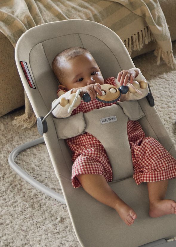 A baby in a red checkered outfit sits in a beige baby bouncer begin, playing with a wooden toy bar Googly eyes. The bouncer is on a soft carpet.