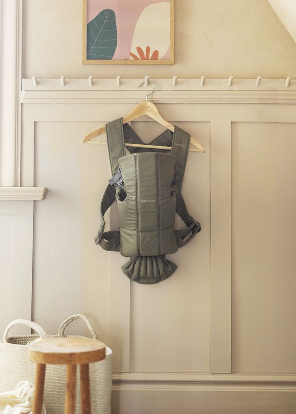 A green baby carrier hangs on a wooden hanger against a beige wall, with a colorful framed picture above and a wooden stool nearby.