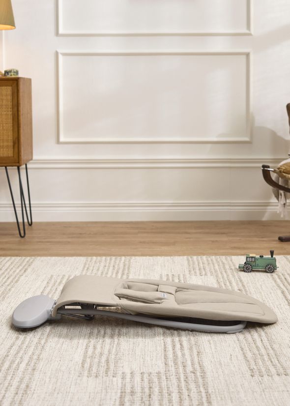 A beige baby bouncer on a striped rug in a cozy room with wooden flooring, vintage cabinet, and a small toy train on the floor.
