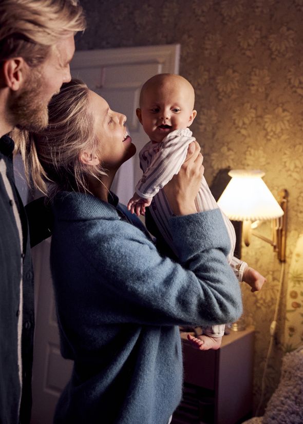 A couple joyfully holds a baby in a warmly lit room with floral wallpaper and a wall lamp.