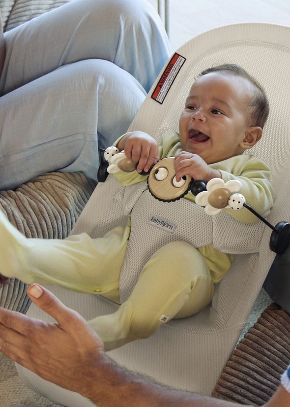 Baby in a bouncer seat, wearing yellow pajamas, smiling joyfully while holding a toy, with an adult's hand nearby.