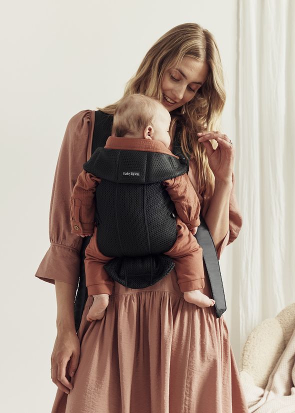 A woman in a pink dress holds a baby in a black carrier. The baby is wearing a rust-colored outfit and is sleeping contently against her chest.