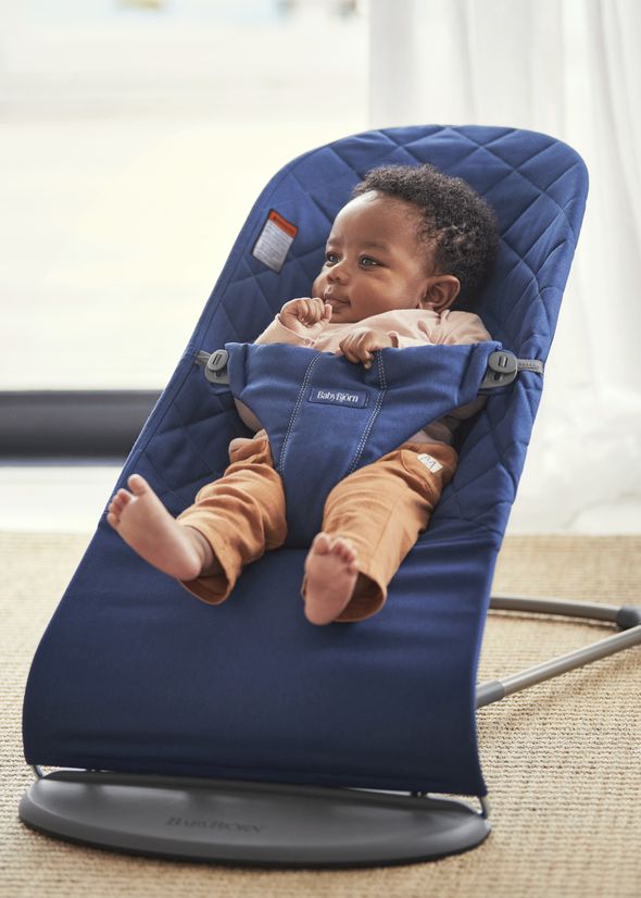 Baby sitting in a blue baby bouncer, wearing a brown outfit, with a calm expression. Bright natural light in the background.