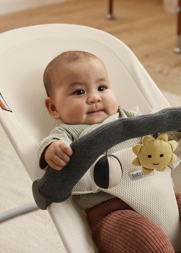 A baby sits in a beige bouncer, holding a gray toy bar with a hanging sun and ball toy, wearing a striped top and red pants, on a wooden floor.