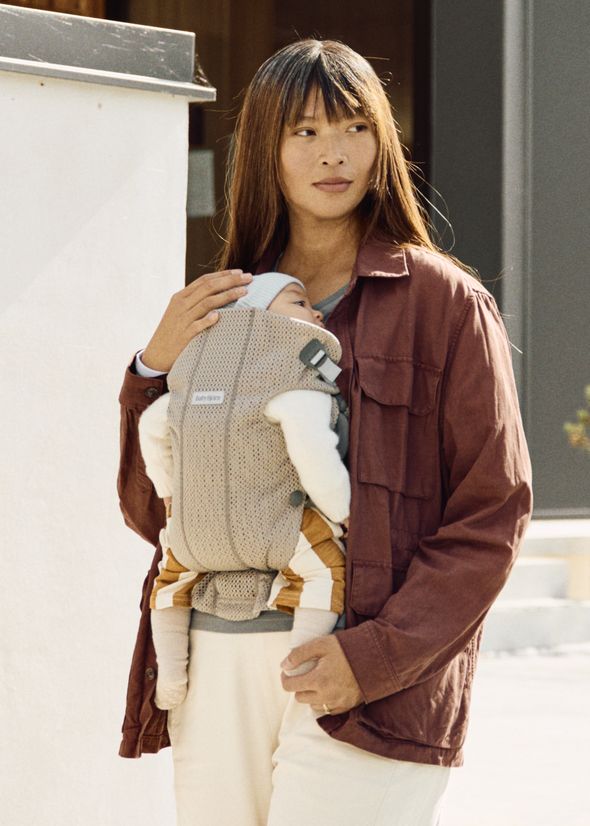 A woman stands outdoors, holding a baby in a beige carrier. She wears a rust-colored jacket and looks to the side.